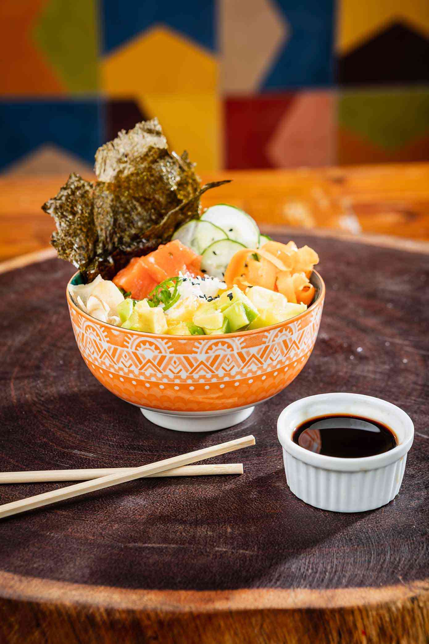 em cima de uma tábua de madeira há um poke, um bowl com arroz, alga, shoio, hachi e salmão
